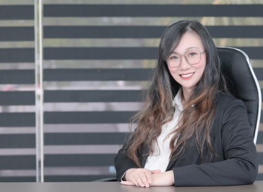 Fraz Joaennie Yeo in dunklem Anzug mit gemusterter Krawatte sitzt an einem Schreibtisch vor einem Fenster mit horizontalen Jalousien.