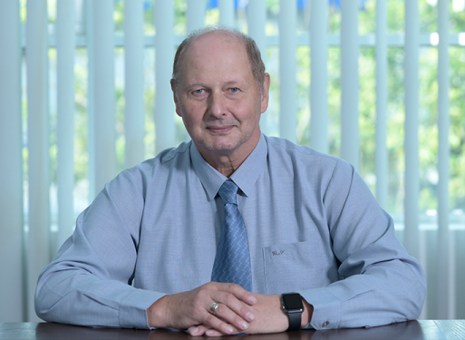 Herr Lee Lih Ren in dunklem Anzug mit gemusterter Krawatte sitzt an einem Schreibtisch vor einem Fenster mit horizontalen Jalousien.