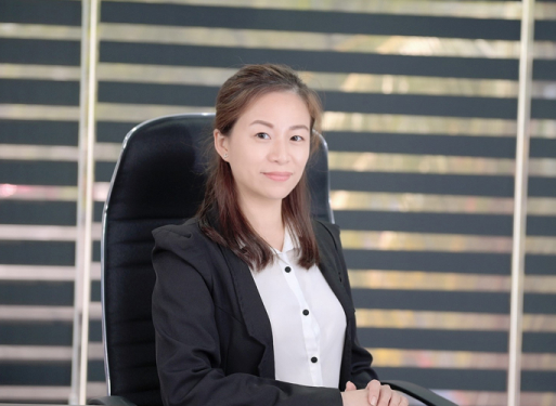 Frau Jenna Liew in dunklem Anzug mit gemusterter Krawatte sitzt an einem Schreibtisch vor einem Fenster mit horizontalen Jalousien.