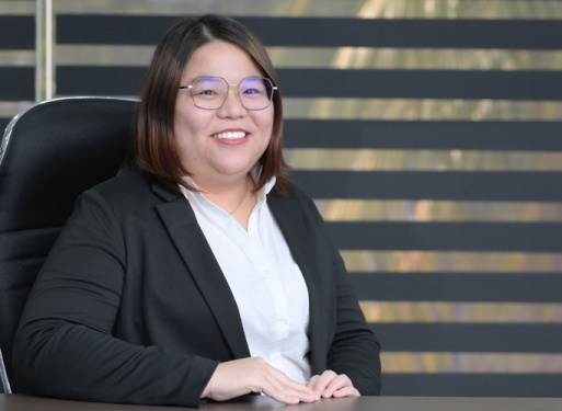 Fraz Joaennie Yeo in dunklem Anzug mit gemusterter Krawatte sitzt an einem Schreibtisch vor einem Fenster mit horizontalen Jalousien.