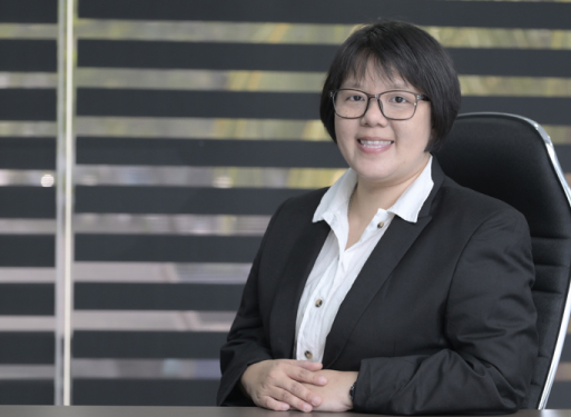 Frau Lam Siau Yen in dunklem Anzug mit gemusterter Krawatte sitzt an einem Schreibtisch vor einem Fenster mit horizontalen Jalousien.