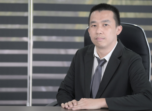 Herr Liow Hock Hinin dunklem Anzug mit gemusterter Krawatte sitzt an einem Schreibtisch vor einem Fenster mit horizontalen Jalousien.