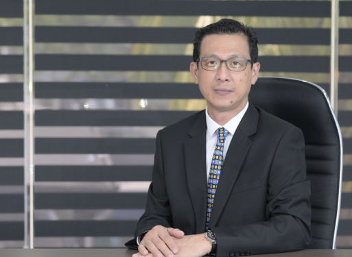 Herr in dunklem Anzug mit gemusterter Krawatte sitzt an einem Schreibtisch vor einem Fenster mit horizontalen Jalousien.