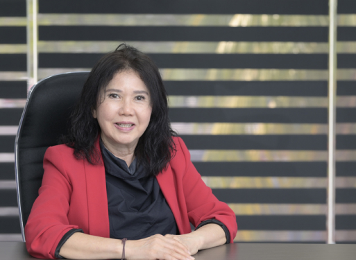 Frau mit dunklem Haar sitzt an einem Tisch, trägt roten Blazer und schwarzes Oberteil, im Hintergrund Jalousien.