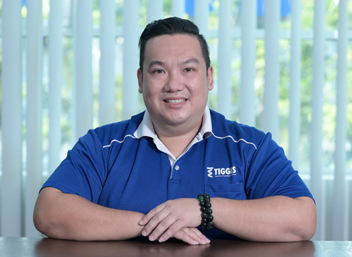 Herr Chong Kok Weng sitzt mit verschränkten Händen an einem Tisch, trägt blaues Poloshirt mit weißem Kragen und Logo auf der Brusttasche.