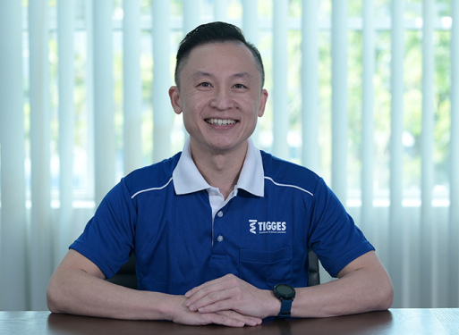 Herr Chow Kah Fai sitzt mit verschränkten Händen an einem Tisch, trägt blaues Poloshirt mit weißem Kragen und Logo auf der Brusttasche.