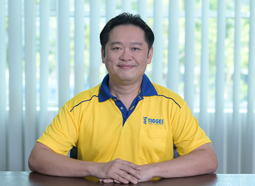 Herr Liew Yuu Seang sitzt mit verschränkten Händen an einem Tisch, trägt gelbes Poloshirt mit blauem Kragen und Logo auf der Brusttasche.