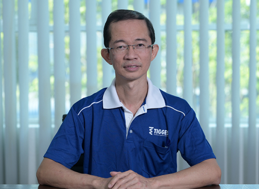 Herr Wong Chee Hoo sitzt mit verschränkten Händen an einem Tisch, trägt blaues Poloshirt mit weißem Kragen und Logo auf der Brusttasche.