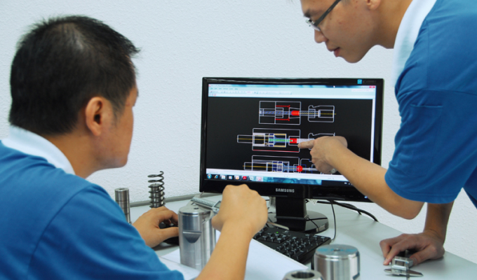 Zwei Männer in blauen Poloshirts betrachten technische Zeichnungen auf einem Computermonitor, einer zeigt mit dem Finger auf den Bildschirm.