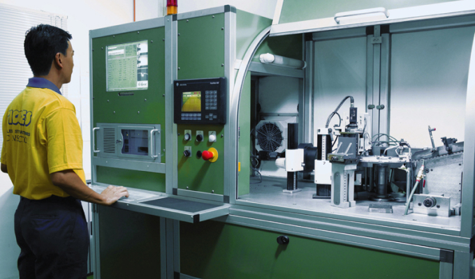 Person steht vor einer grünen Industriemaschine mit Monitoren und Bedienelementen in einem Labor oder Werkstatt.