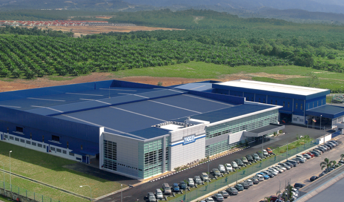 Außenansicht des TIGGES-Standorts in Ipoh: modernes Produktions- und Verwaltungsgebäude mit Glasfassade, Parkplatz im Vordergrund und bewaldeter Hügellandschaft im Hintergrund, aufgenommen bei Tageslicht.