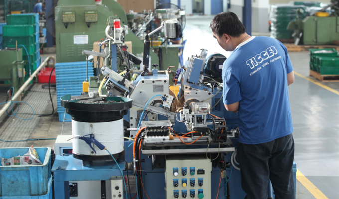 Mann in blauem T-Shirt mit Aufschrift "TIGGES" arbeitet an komplexer Maschine in Industriehalle.