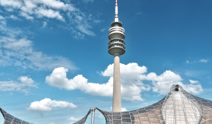 Fernsehturm mit rundem Aussichtsbereich vor einer gewölbten, netzartigen Dachkonstruktion unter blauem Himmel mit Wolken.