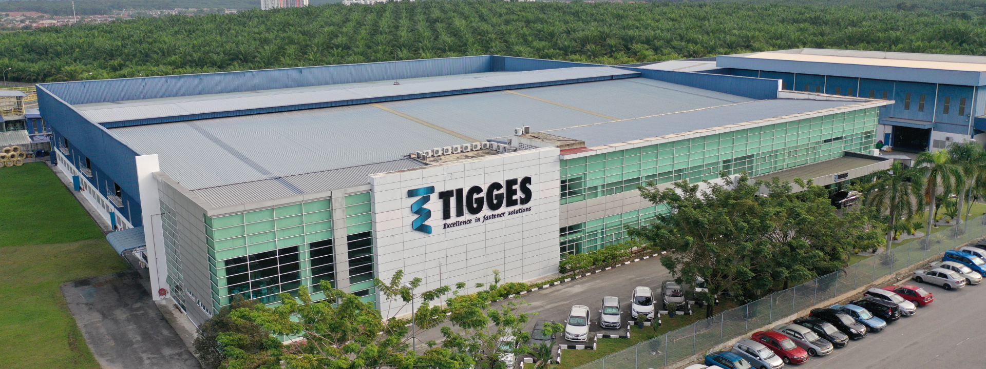 Außenansicht des TIGGES-Standorts in Ipoh: modernes Produktions- und Verwaltungsgebäude mit Glasfassade, Parkplatz im Vordergrund und bewaldeter Hügellandschaft im Hintergrund, aufgenommen bei Tageslicht.