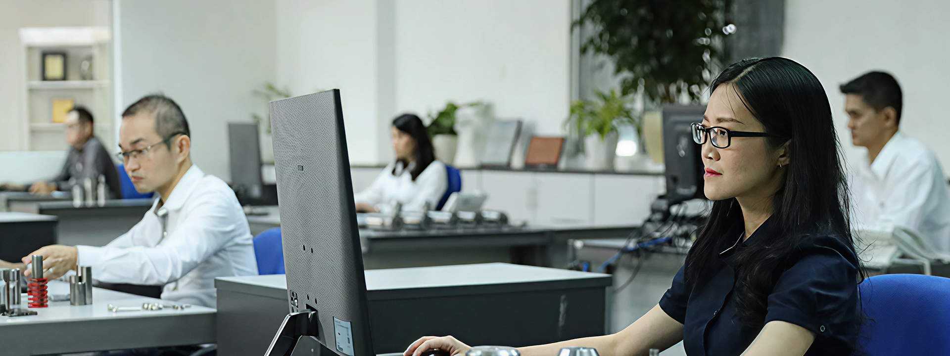 Mitarbeitende in einem modernen Büro- und Entwicklungsumfeld; im Vordergrund arbeitet eine Person am Computer, im Hintergrund weitere Beschäftigte an technischen Bauteilen