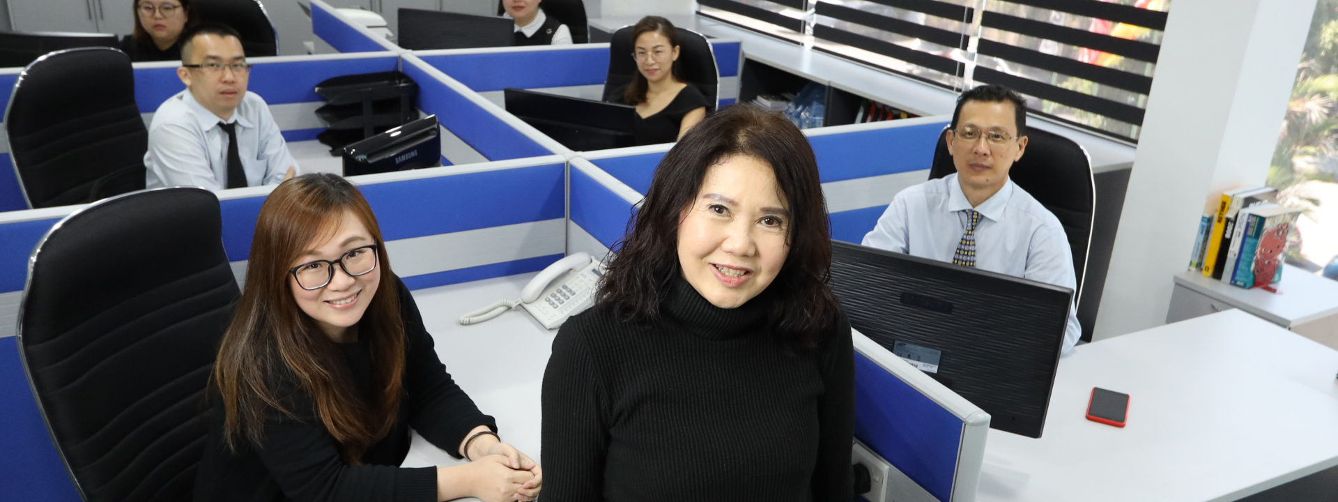 Mehrere Personen sitzen an Schreibtischen in einem Büro mit blauen Trennwänden und Computern.