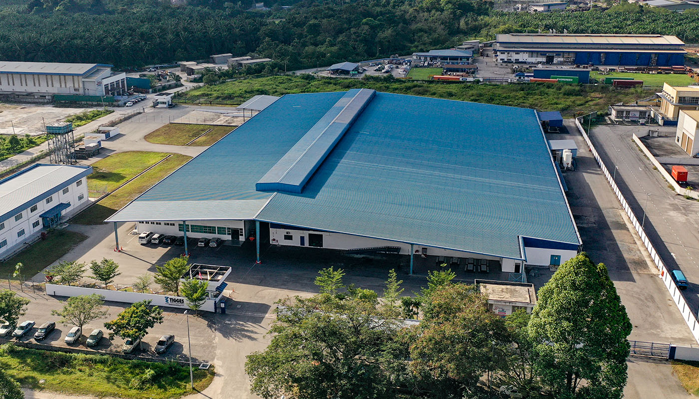 Luftaufnahme eines TIGGES-Produktionsstandorts mit großer Industriehalle, Ladebereich und umgebender Infrastruktur in grüner Umgebung.