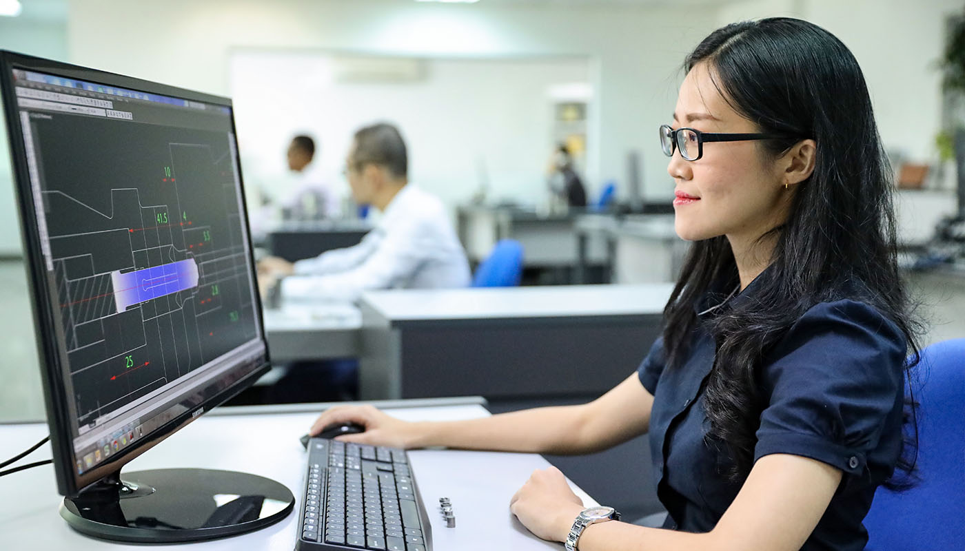 Mitarbeiterin im technischen Büro arbeitet am Computer mit CAD-Software zur Konstruktion von Bauteilen, im Hintergrund weitere Arbeitsplätze.