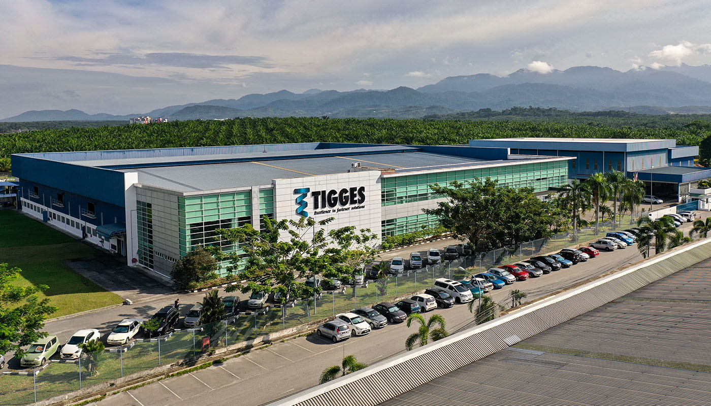 Luftaufnahme des TIGGES-Produktions- und Verwaltungsgebäudes mit Parkplatz, umgeben von Grünflächen und Bergen im Hintergrund.